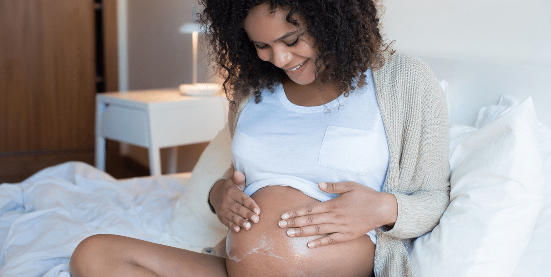 Pregnant woman sitting on a bed rubbing lotion on her belly